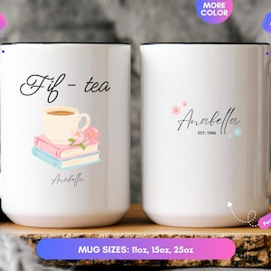 May include: A white ceramic mug with a black handle and rim. The front features the text 'Fif-tea' above an illustration of a teacup on two books and the name 'Anabella'. The back displays the name 'Anabella' with 'EST: 1986' and small floral accents.