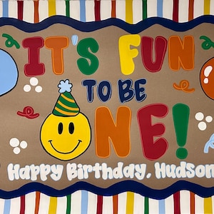 May include: A rectangular birthday sign with a striped border and a tan center featuring the text IT'S FUN TO BE ONE! and Happy Birthday, Hudson! A yellow smiley face wearing a party hat, green, blue, orange, and red balloons, and confetti.