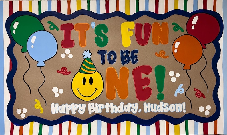 May include: A rectangular birthday sign with a striped border and a tan center featuring the text IT'S FUN TO BE ONE! and Happy Birthday, Hudson! A yellow smiley face wearing a party hat, green, blue, orange, and red balloons, and confetti.
