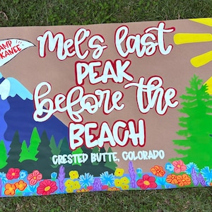 May include: A rectangular banner featuring a hand-painted mountain landscape with a blue peak, a white flag labeled Camp Kokanee, green pine trees, a yellow sun, and colorful wildflowers. The text reads Mel's last PEAK before the BEACH CRESTED BUTTE, COLORADO.