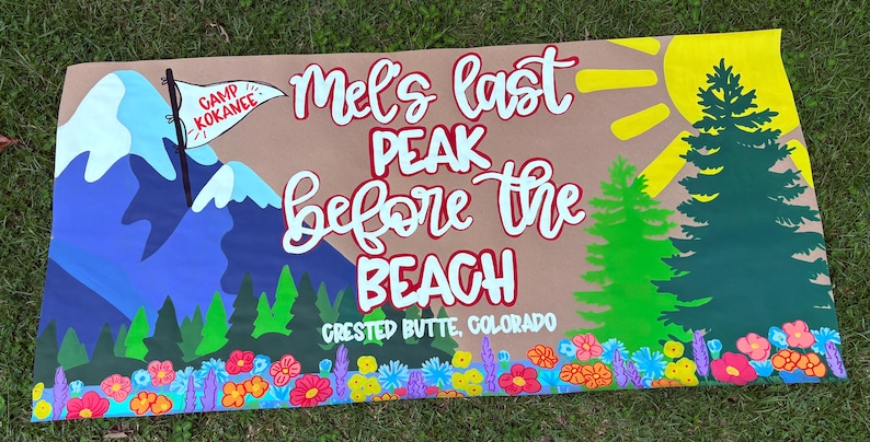 May include: A rectangular banner featuring a hand-painted mountain landscape with a blue peak, a white flag labeled Camp Kokanee, green pine trees, a yellow sun, and colorful wildflowers. The text reads Mel's last PEAK before the BEACH CRESTED BUTTE, COLORADO.