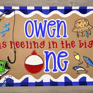 May include: A rectangular banner with a blue and white buffalo check border surrounding a tan center. The text reads "OWEN is reeling in the big one" in blue and red. Illustrations include a fishing rod, a green fish, a hat, and lures.