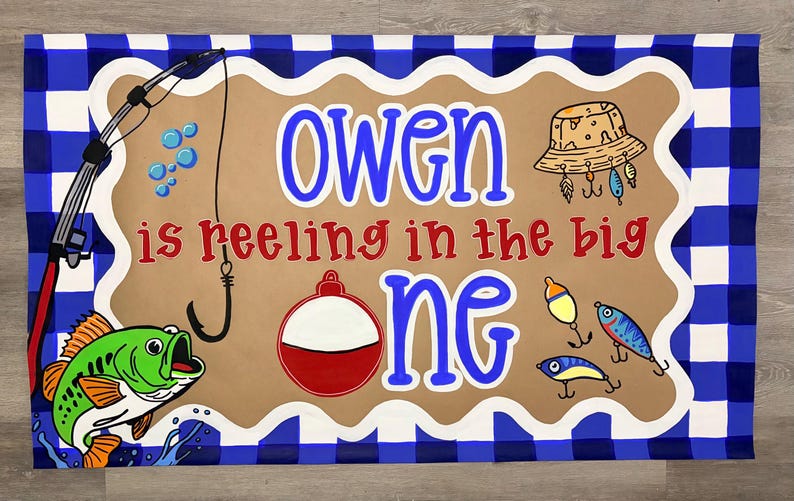 May include: A rectangular banner with a blue and white buffalo check border surrounding a tan center. The text reads "OWEN is reeling in the big one" in blue and red. Illustrations include a fishing rod, a green fish, a hat, and lures.