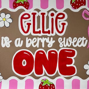 May include: A rectangular sign with a pink and white striped border featuring red strawberries and white flowers. The center is brown with the text GLLIG is a berry sweet ONE in red and white lettering, accented by a pink bow on the right side.