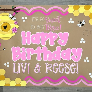 May include: A rectangular brown poster featuring a yellow beehive with pink flowers, yellow honeycomb patterns, and cartoon bees. The text reads: It's so sweet to bee three! Happy Birthday Livi & Reese! The design includes pink wavy border lines.