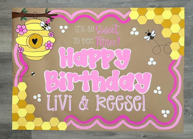 May include: A rectangular brown poster featuring a yellow beehive with pink flowers, yellow honeycomb patterns, and cartoon bees. The text reads: It's so sweet to bee three! Happy Birthday Livi & Reese! The design includes pink wavy border lines.