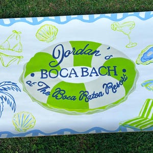 May include: A rectangular beach towel featuring a light blue and white striped border. The center displays a green and white striped pool float with the text "Jordan's BOCA BACH at The Boca Raton Resort" surrounded by illustrations of a palm tree, shells, a bikini, a cocktail, and a lounge chair.
