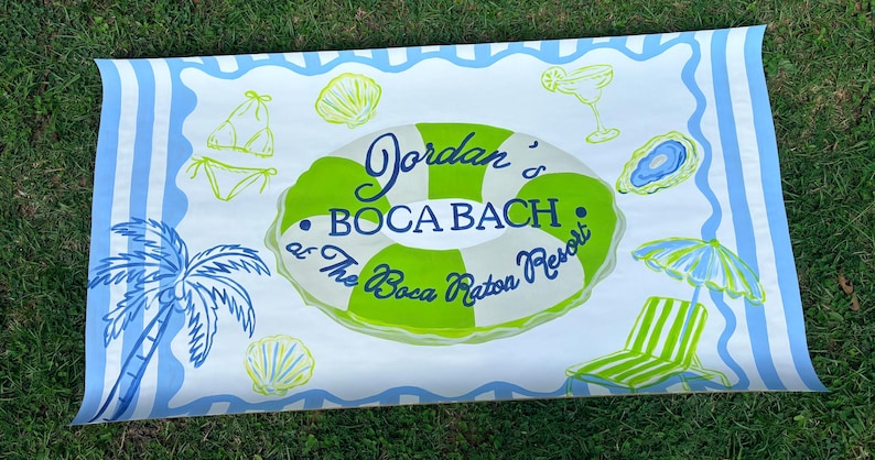 May include: A rectangular beach towel featuring a light blue and white striped border. The center displays a green and white striped pool float with the text "Jordan's BOCA BACH at The Boca Raton Resort" surrounded by illustrations of a palm tree, shells, a bikini, a cocktail, and a lounge chair.