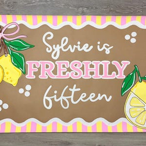 May include: A rectangular banner featuring a yellow and pink vertical striped border. The center is a brown scalloped rectangle with the white and pink text Sylvie is FRESHLY fifteen, decorated with yellow lemons, green leaves, and a pink bow.