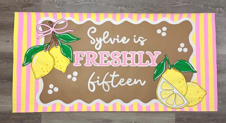 May include: A rectangular banner featuring a yellow and pink vertical striped border. The center is a brown scalloped rectangle with the white and pink text Sylvie is FRESHLY fifteen, decorated with yellow lemons, green leaves, and a pink bow.