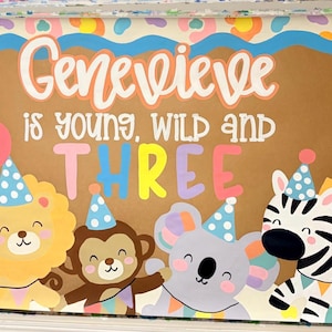 May include: A rectangular sign featuring the text Genevieve is young, wild and THREE in white and colorful lettering. The design includes illustrations of a blue dinosaur, yellow lion, brown monkey, gray koala, zebra, and giraffe wearing party hats.