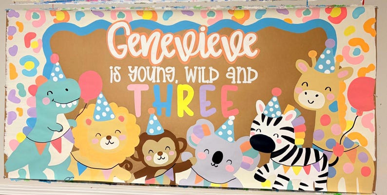 May include: A rectangular sign featuring the text Genevieve is young, wild and THREE in white and colorful lettering. The design includes illustrations of a blue dinosaur, yellow lion, brown monkey, gray koala, zebra, and giraffe wearing party hats.