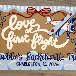 May include: A rectangular sign with a tan center and light blue striped border. It features a red bow, a small airplane, a diamond ring, and hearts. The text reads: Love at first flight, Maddie's Bachelorette Trip, and CHARLESTON, SC 2026.