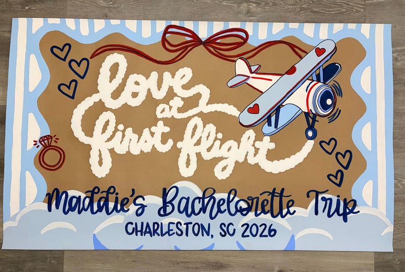 May include: A rectangular sign with a tan center and light blue striped border. It features a red bow, a small airplane, a diamond ring, and hearts. The text reads: Love at first flight, Maddie's Bachelorette Trip, and CHARLESTON, SC 2026.