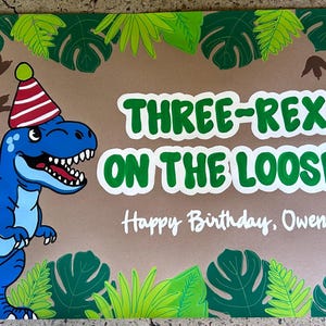 May include: A rectangular sign featuring a blue cartoon dinosaur wearing a red and white striped party hat. The text reads "THREE-REX ON THE LOOSE!" and "Happy Birthday, Owen!" The border is decorated with green tropical leaves and brown dinosaur footprints.