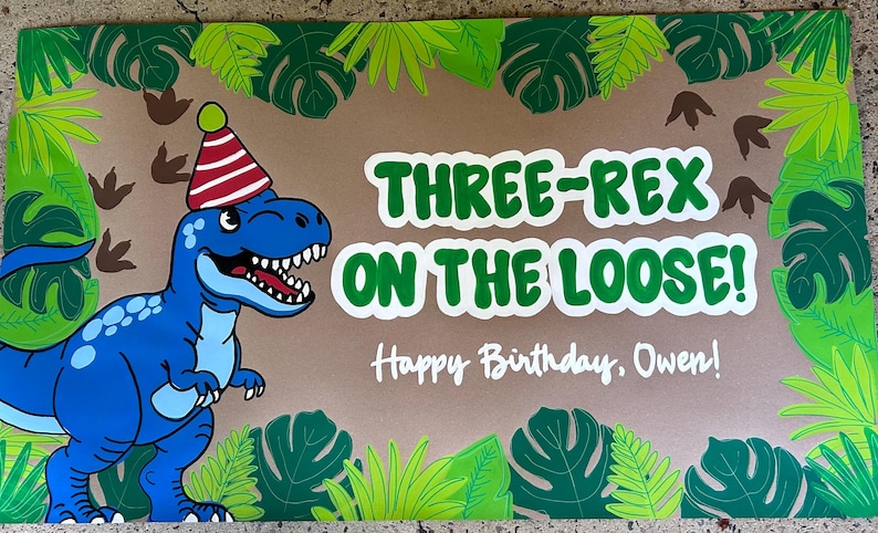 May include: A rectangular sign featuring a blue cartoon dinosaur wearing a red and white striped party hat. The text reads "THREE-REX ON THE LOOSE!" and "Happy Birthday, Owen!" The border is decorated with green tropical leaves and brown dinosaur footprints.