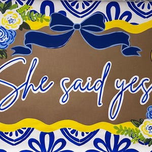 May include: A rectangular sign featuring the text "She said yes!" in white script with a blue outline. The sign has a brown center, a yellow wavy border, and a blue and white tile pattern background. It is decorated with blue and yellow flowers, a dark blue bow, and a gold engagement ring illustration.