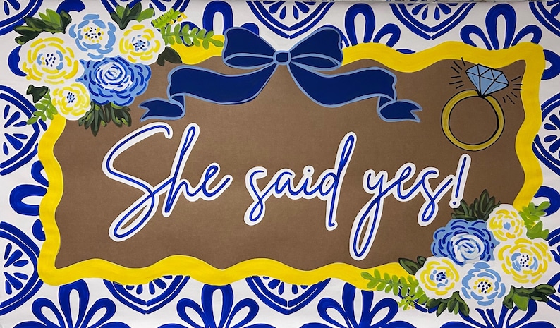 May include: A rectangular sign featuring the text "She said yes!" in white script with a blue outline. The sign has a brown center, a yellow wavy border, and a blue and white tile pattern background. It is decorated with blue and yellow flowers, a dark blue bow, and a gold engagement ring illustration.