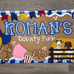 May include: A rectangular banner with a blue and white checkered border featuring the text ROMAN'S County Fair. Illustrations include a brown pony, pink cotton candy, popcorn, a pretzel, three goats behind a fence, balloons, and a blue ribbon with the number 4.