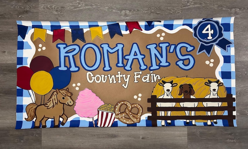May include: A rectangular banner with a blue and white checkered border featuring the text ROMAN'S County Fair. Illustrations include a brown pony, pink cotton candy, popcorn, a pretzel, three goats behind a fence, balloons, and a blue ribbon with the number 4.
