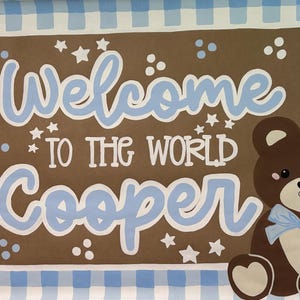 May include: A rectangular sign with a blue and white gingham border surrounding a brown center. The text reads Welcome TO THE WORLD Cooper in light blue and white lettering. A brown teddy bear with a light blue bow sits on the right side.