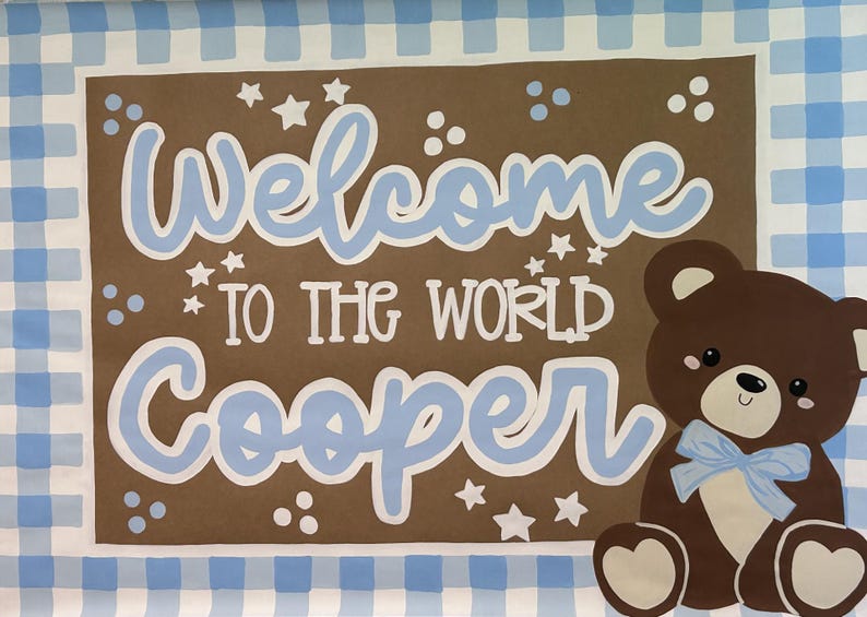 May include: A rectangular sign with a blue and white gingham border surrounding a brown center. The text reads Welcome TO THE WORLD Cooper in light blue and white lettering. A brown teddy bear with a light blue bow sits on the right side.