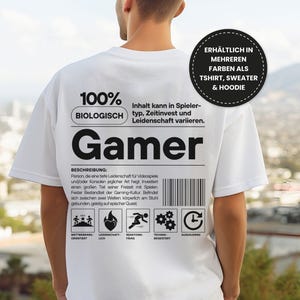 May include: A white t-shirt with the word "Gamer" in large black letters. The shirt also features text in German, including "100% BIOLOGISCH" and a description of gamers. A circular graphic states the shirt is available in multiple colors, as a t-shirt, sweater, and hoodie.