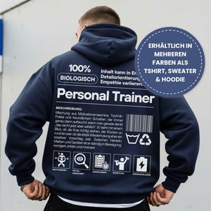 Könnte beinhalten: Marineblauer Hoodie mit weißem Text und Grafiken auf der Rückseite, darunter "Personal Trainer" und eine Beschreibung. Der Hoodie hat auch eine runde Grafik mit der Aufschrift "Erhältlich in mehreren Farben als T-Shirt, Sweater & Hoodie."