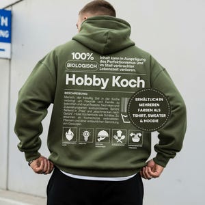 Op de afbeelding: Een olijfgroene hoodie met de Duitse tekst "Hobby Koch" op de rug. De hoodie heeft een capuchon en een witte afbeelding met tekst en iconen. Verkrijgbaar als t-shirt, trui en hoodie.