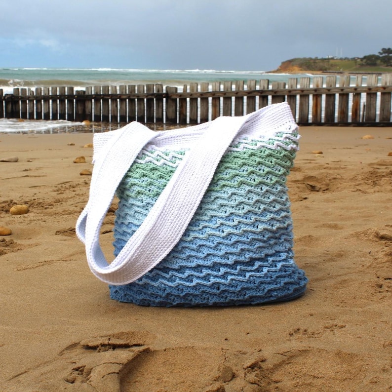 May include: A handmade crochet tote bag in shades of blue and green, with white straps. The bag is sitting on a sandy beach with the ocean in the background. The bag's design features a chevron pattern.