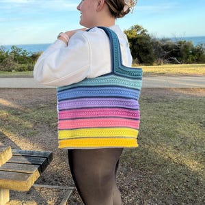 May include: A large, handmade crochet tote bag with a vibrant rainbow gradient design. The bag transitions from yellow to pink, purple, blue, and teal. The handles and top border are a darker teal. The bag is carried over the shoulder.