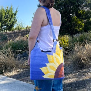 Peut inclure: Un sac fourre-tout en crochet bleu avec un motif de tournesol. Le sac a une bandoulière bleue et des poignées en anneaux de bois. Le tournesol est jaune, marron et violet. Le sac est porté à l'épaule.