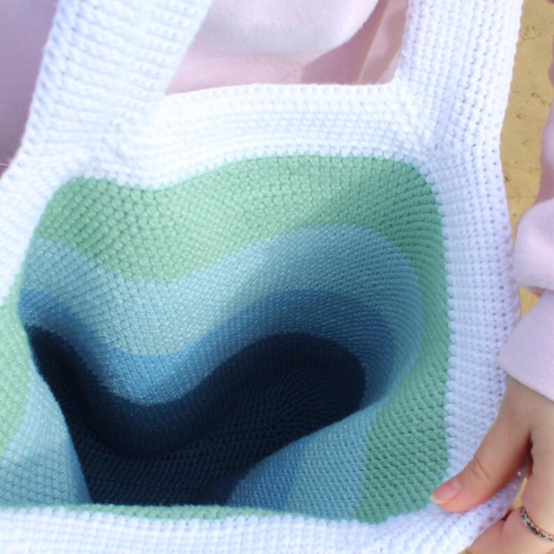May include: A handmade crochet tote bag with a white border and handles. The interior features a gradient of green, blue, and navy blue. The bag is made of a textured yarn, and the open top is visible.