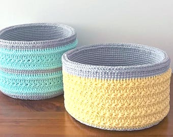 May include: Two handmade crochet baskets. One is turquoise and gray, the other is yellow and gray. The baskets are cylindrical with a textured stitch pattern. The baskets are sitting on a wooden surface.