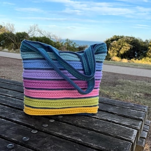 May include: A handmade crochet tote bag with a striped pattern in shades of blue, purple, pink, yellow, and green. The bag has two handles and is sitting on a wooden picnic table. The background shows a view of the ocean and sky.