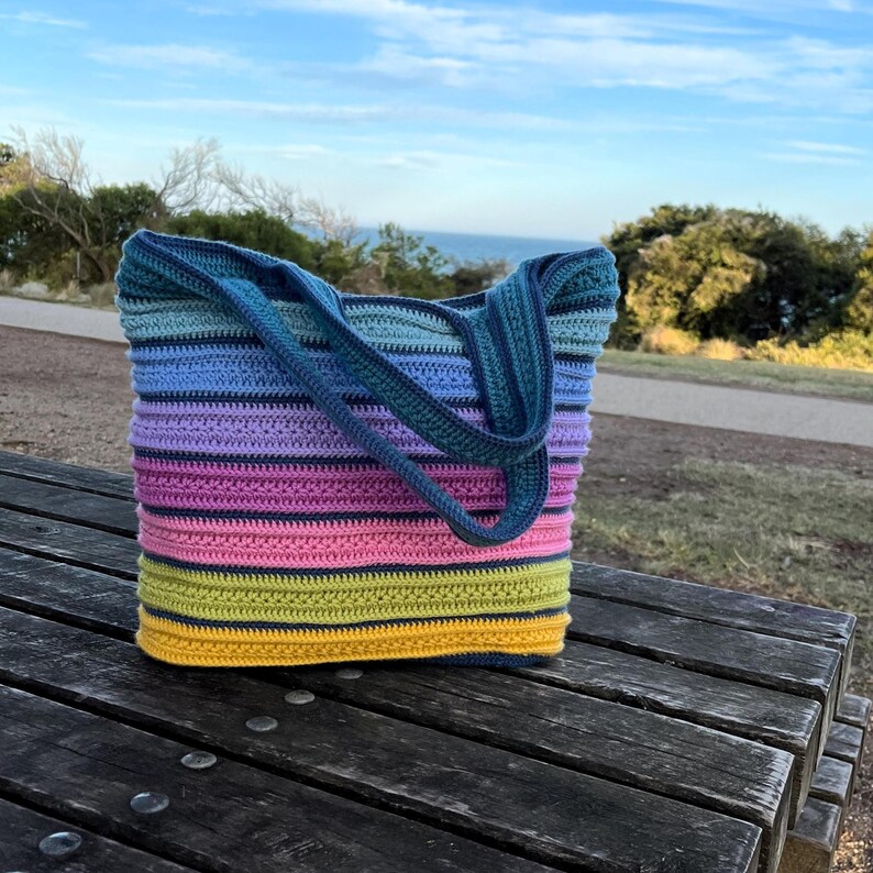 May include: A handmade crochet tote bag with a striped pattern in shades of blue, purple, pink, yellow, and green. The bag has two handles and is sitting on a wooden picnic table. The background shows a view of the ocean and sky.