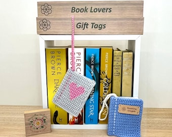Puede incluir: Una estantería blanca exhibe libros y marcadores de ganchillo. Un marcador gris tiene un corazón rosa, y un marcador azul tiene una etiqueta Blooming Delights. Los letreros de madera de arriba dicen "Book Lovers" y "Gift Tags".