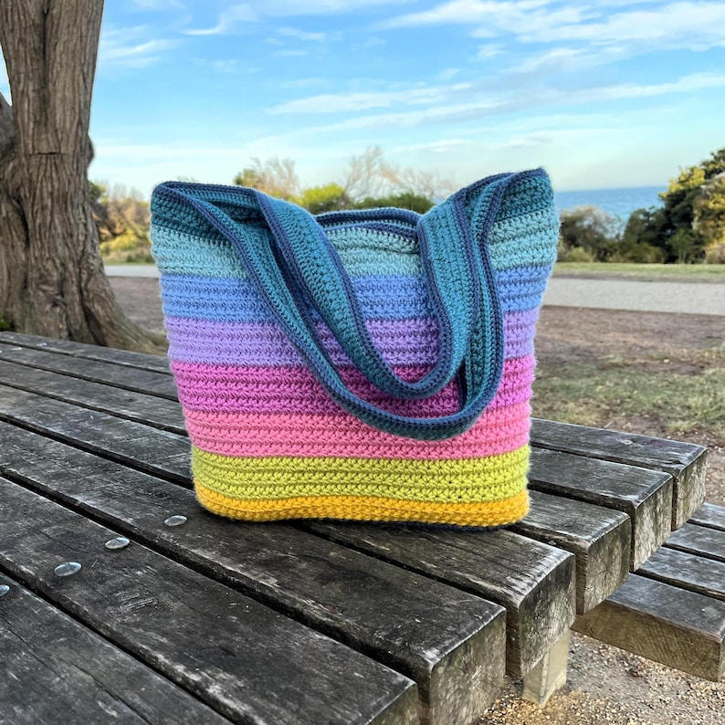 May include: A handmade crochet tote bag with a vibrant rainbow color scheme. The bag features horizontal stripes of yellow, pink, purple, blue, and teal. The handles and top trim are a darker teal color. The bag is sitting on a wooden picnic table.