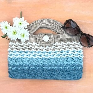 May include: A crocheted handbag in a blue, white, and grey wave design. The bag features a rounded handle and a small flap secured with a white button. Sunglasses and white flowers are placed beside the bag.