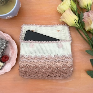 Könnte beinhalten: Eine gehäkelte Handytasche mit einem Farbverlauf in Rosa, Weiß und Beige. Die Tasche hat einen gewellten Rand und ein kleines Fach für ein Handy. Die Tasche liegt auf einer Holzoberfläche.