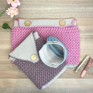 May include: A collection of handmade crochet items in shades of pink, gray, and purple. The set includes a large pouch with wooden buttons, a small pouch, and a small container with blue stripes. The items are displayed on a wooden surface.