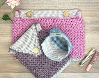 May include: A selection of handmade crochet items in pink, grey, and purple. The set includes a large pouch with wooden buttons, a small pouch, and a small container with blue stripes. The items are displayed on a wooden surface.