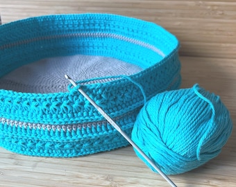 May include: A turquoise crochet basket with a grey trim is being worked on with a crochet hook and a ball of turquoise yarn. The basket is sitting on a wooden surface.