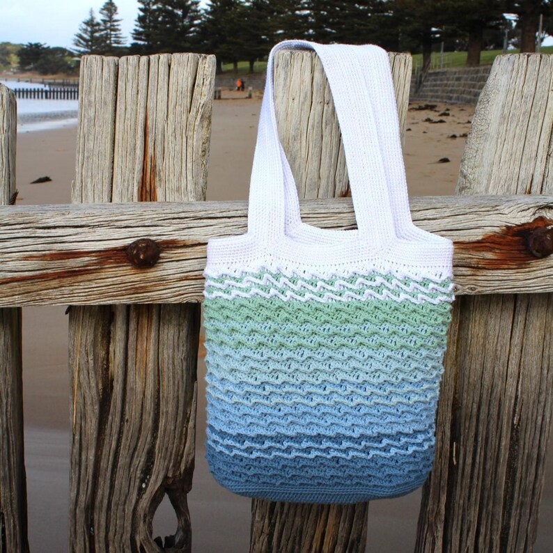 May include: A white and blue crochet tote bag with a wave pattern. The bag has white handles and a gradient of colors, transitioning from green to blue. The bag is hanging on a weathered wooden fence.