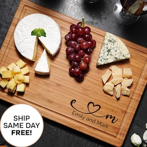 Könnte beinhalten: Ein hölzernes Charcuterie-Brett mit einer Nut am Rand, mit Käse, Trauben und einem Herzdesign mit den Namen "Emily and Mark". Das Brett ist von Weingläsern, Kerzen und Crackern umgeben.