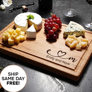 Puede incluir: Una tabla de embutidos de madera con varios quesos, uvas rojas y los nombres "Emily and Mark" grabados con un corazón. Copas de vino, velas y una botella de vino están dispuestas alrededor.