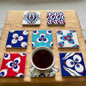 Op de afbeelding: Een verzameling van acht vierkante keramische onderzetters met diverse traditionele patronen in rood, blauw, wit en turquoise. Eén onderzetter ligt onder een witte keramische kop gevuld met donkere thee, terwijl de andere eromheen zijn gerangschikt.