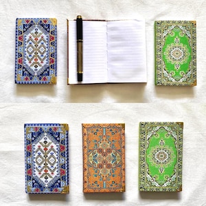 May include: A collection of small notebooks and a journal with covers patterned like oriental rugs. The notebooks have blue, green, and orange designs with gold corner accents. A pen rests on an open journal with lined pages.