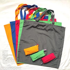 Pode incluir: Uma coleção de sacolas coloridas em laranja, azul, vermelho, verde e cinza. Cada sacola tem alças combinando. Três bolsas dobradas com zíper em laranja, verde e vermelho estão na frente da sacola cinza. As sacolas são feitas de um material leve.