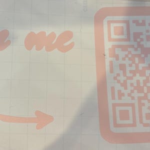 May include: A sticker featuring the text "Scan me" in a cursive font, with an arrow pointing to a QR code. The text and QR code are in a pale pink hue.